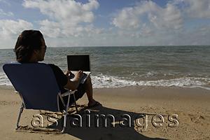 Asia Images Group - Rear view of man sitting on beach working on a laptop