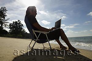 Asia Images Group - Man sitting on beach chair working on laptop at the beach