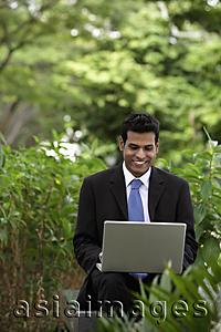 Asia Images Group - Indian man working on laptop with green plants and trees in foreground and background