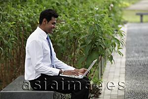 Asia Images Group - Indian man sitting outside working on laptop with plants in background