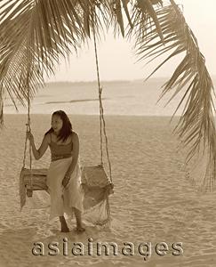 Asia Images Group - Woman sitting on swing under coconut tree looking off camera, sea in background