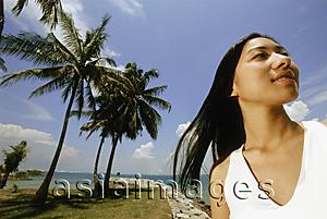 Asia Images Group - Young woman standing on beach, looking up, coconut trees in background, low angle view