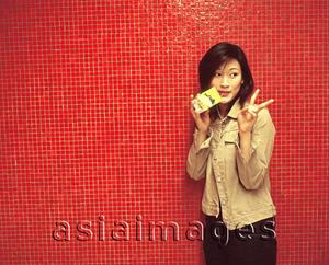 Asia Images Group - Young woman making a peace sign, drinking a packet drink, red background.
