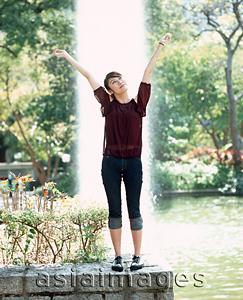Asia Images Group - Young woman standing with arms outstretched, fountain in background.