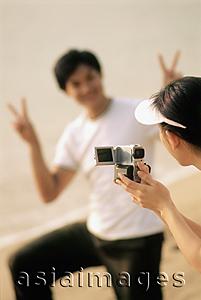 Asia Images Group - Couple at beach, woman filming man making peace sign