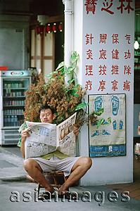 Asia Images Group - Singapore, Chinatown, Man sitting on stool reading local Chinese newspaper.