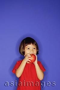 Asia Images Group - Young girl standing against blue background, eating an apple