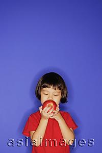 Asia Images Group - Young girl standing against blue background, holding an apple and biting it.