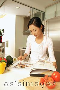 Asia Images Group - Young woman standing at kitchen counter, looking at cookbook