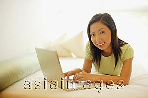 Asia Images Group - Young woman lying on bed, using laptop, looking at camera