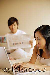 Asia Images Group - Couple in bedroom, man reading newspaper, woman using laptop