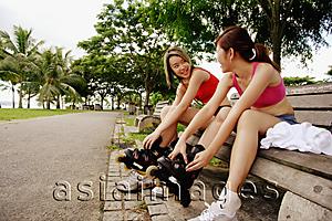 Asia Images Group - Two women sitting side by side on park bench, putting on roller blades