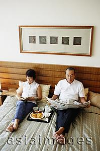 Asia Images Group - Older couple lying on bed, reading newspaper, tray of food between them
