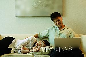 Asia Images Group - Man sitting on sofa, using laptop, woman lying next to him