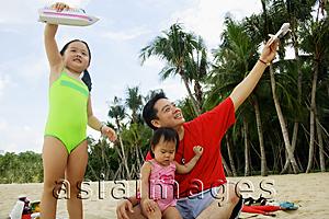 Asia Images Group -  Father with two girls, playing on beach with toy boat and toy plane