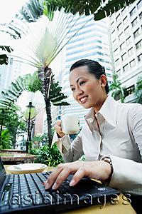 Asia Images Group - Business woman using laptop, holding coffee cup