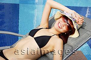 Asia Images Group -  Young woman in bikini, wearing hat and sunglasses, lounging by pool, looking at camera
