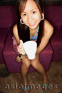 Asia Images Group - Young woman sitting on sofa, holding cup, looking up at camera