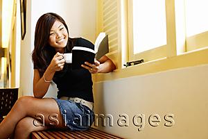 Asia Images Group - Young woman sitting next to window, reading a book and holding cup of coffee