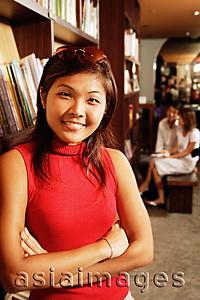 Asia Images Group - Young woman leaning on book shelf, arms crossed, looking at camera