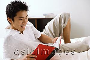 Asia Images Group - Man lying on bed, reading book, smiling