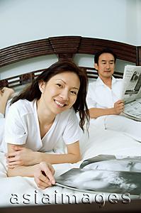 Asia Images Group - Couple in bedroom, woman smiling at camera, man reading newspaper in the background