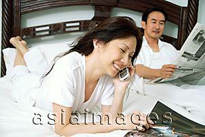 Asia Images Group - Couple in bedroom, woman reading magazine and using mobile phone, man reading newspaper