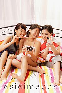 Asia Images Group - Three young women sitting on bed, side by side, applying make-up
