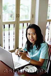 Asia Images Group - Woman using laptop, holding cup, looking at camera