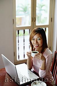 Asia Images Group - Woman using laptop, holding cup, smiling at camera