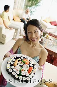 Asia Images Group - Woman holding up plate of sushi, looking at camera, people in the background