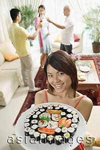 Asia Images Group - Woman holding up plate of sushi, smiling at camera, people in the background