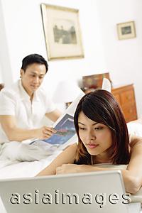 Asia Images Group - Couple in bedroom, woman lying on bed, looking at laptop, man reading newspaper behind her