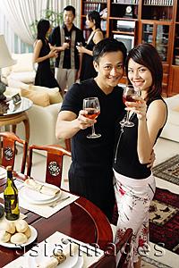 Asia Images Group - Couple in living room, raising wine glasses to camera, people in the background