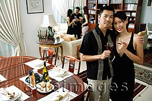 Asia Images Group - Couple at home, holding wine glasses, smiling at camera, people in the background