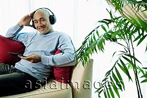 Asia Images Group - Man on sofa with headphones, listening to music, smiling at camera
