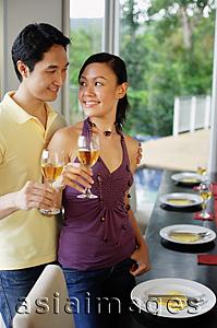 Asia Images Group - Couple standing next to dining table, holding wine glasses, looking at each other