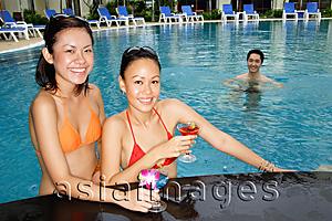Asia Images Group - Young women in swimming pool, holding cocktails, smiling at camera,  man swimming behind them