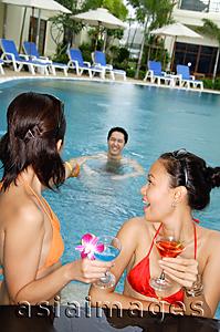 Asia Images Group - Young women in swimming pool, holding cocktails, looking at man swimming behind them