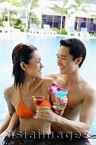 Asia Images Group - Couple in swimming pool holding cocktail drinks, smiling at each other