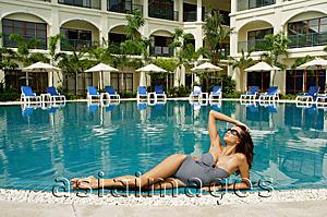 Asia Images Group - Woman lying next to swimming pool, hand on head, building in the background