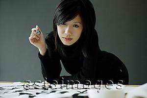 Asia Images Group - Woman holding paintbrush, leaning on table, looking at camera