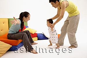 Asia Images Group - Mother sitting on sofa, arms outstretched, father helping toddler walk towards her