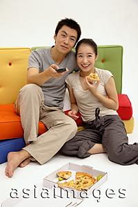 Asia Images Group - Couple sitting on sofa, woman eating pizza, man holding TV remote control