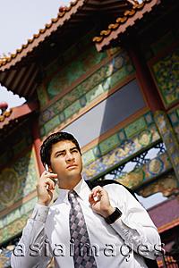 Asia Images Group - Businessman standing in front of temple gate, holding jacket over shoulder, using mobile phone