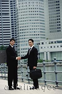 Asia Images Group - Two businessmen shaking hands, looking at camera, river and buildings in the background