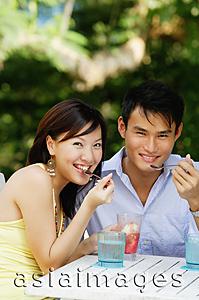 Asia Images Group - Couple sitting side by side, eating, smiling at camera