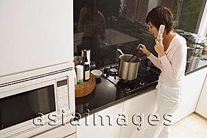 Asia Images Group - Young woman cooking in kitchen, using mobile phone