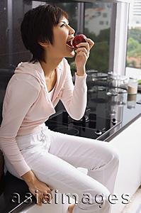 Asia Images Group - Young woman sitting on kitchen counter, eating an apple