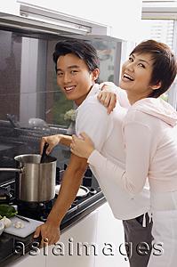 Asia Images Group - Man cooking on stove, woman embracing him from behind, both looking at camera